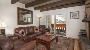 A living room with a brown leather sofa, a wooden coffee table, and a patterned rug. There is a large mirror on the wall and a framed picture above a fireplace. - Meadow Ridge Townhome A (Vail)