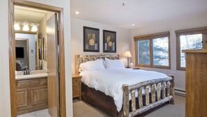 Un dormitorio con cama de madera, una mesita de noche, una lámpara y un aparador. Hay un espejo grande sobre el aparador y dos cuadros en la pared.