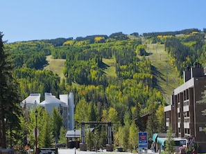 A building with a sign for Lionshead Village, a parking area, and a view of a forested hillside. - Vantage Point Condo 105 (Vail)