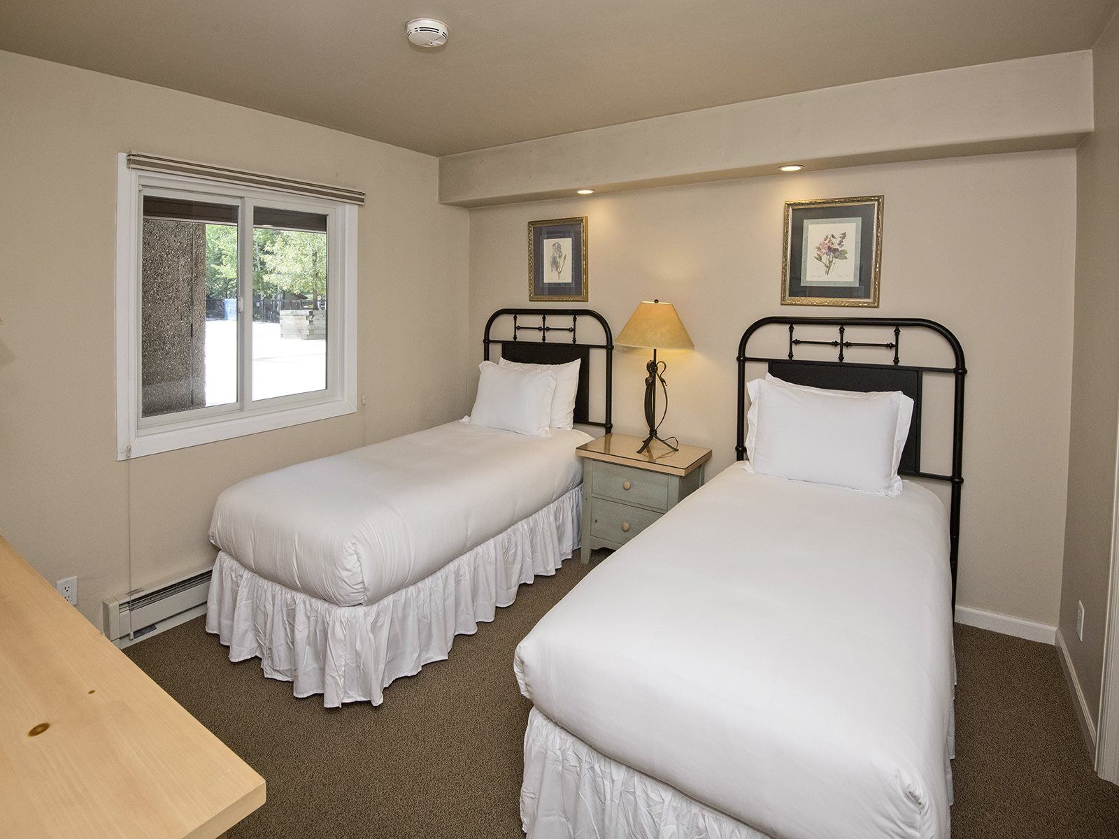 A twin room with two single beds, a small bedside table with a lamp, a window with blinds, and a picture frame on the wall.