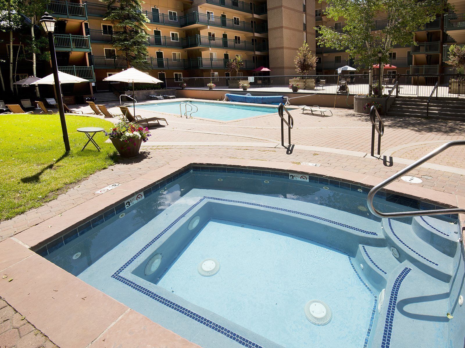 A rectangular pool with a tiled border, surrounded by lounge chairs and umbrellas, with a multi-story building in the background.