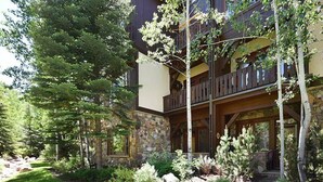 A multi-story building with a stone facade, wooden balconies, and a well-maintained garden with various plants and trees. - Arrowhead Village Condo - 104 Aspenwood Lodge (Edwards)