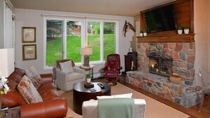 A living room with a stone fireplace, leather sofas, a TV mounted above it, and a view of a green lawn through large windows. - Arrowhead Village Condo - 104 Seasons Lodge (Edwards)