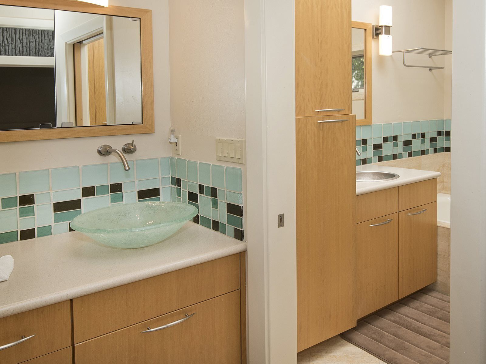 Un baño con lavamanos de cristal, armarios de madera, un espejo y salpicadero de azulejos.