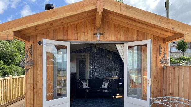 Una cabaña de madera con la puerta abierta, mostrando un interior acogedor con un sofá y una zona de comedor.