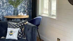 Una sala de estar con un sofá azul oscuro, una mesa de centro de vidrio, un sillón azul, una mesita de madera con un jarrón y una tetera, y un ventanal con vistas a la vegetación.