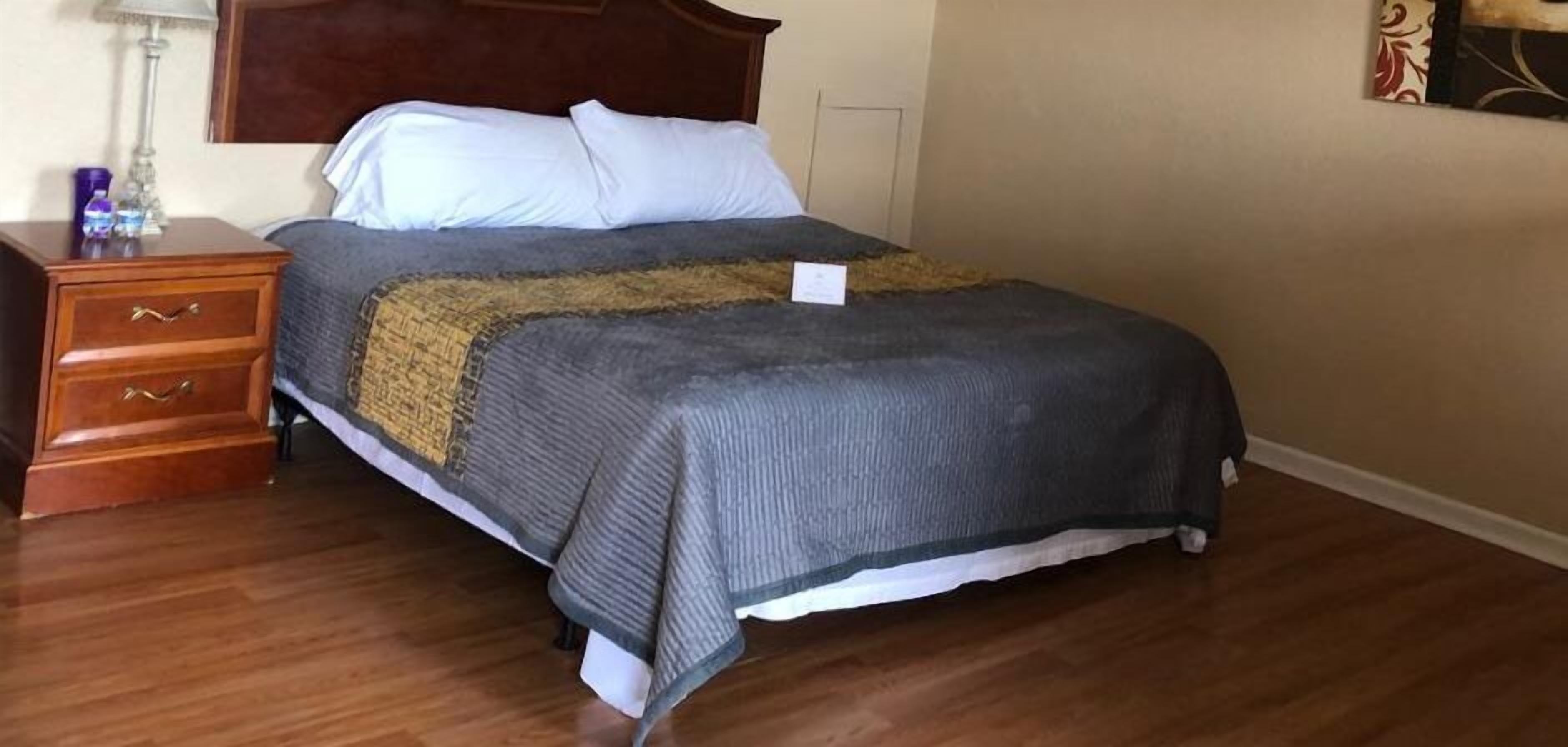 Un lit bien fait, recouvert d’une couverture grise et beige, une table de chevet en bois avec des tiroirs et une lampe.