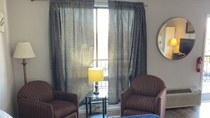 Ein Hotelzimmer mit einem großen Bett, zwei Sesseln, einem kleinen Tisch mit Lampe, einem runden Spiegel und einem Fenster mit Vorhängen.