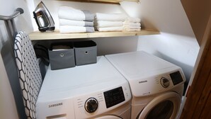 Un espace buanderie équipé de deux machines à laver Samsung, d’une étagère avec des serviettes pliées et d’un fer à repasser.