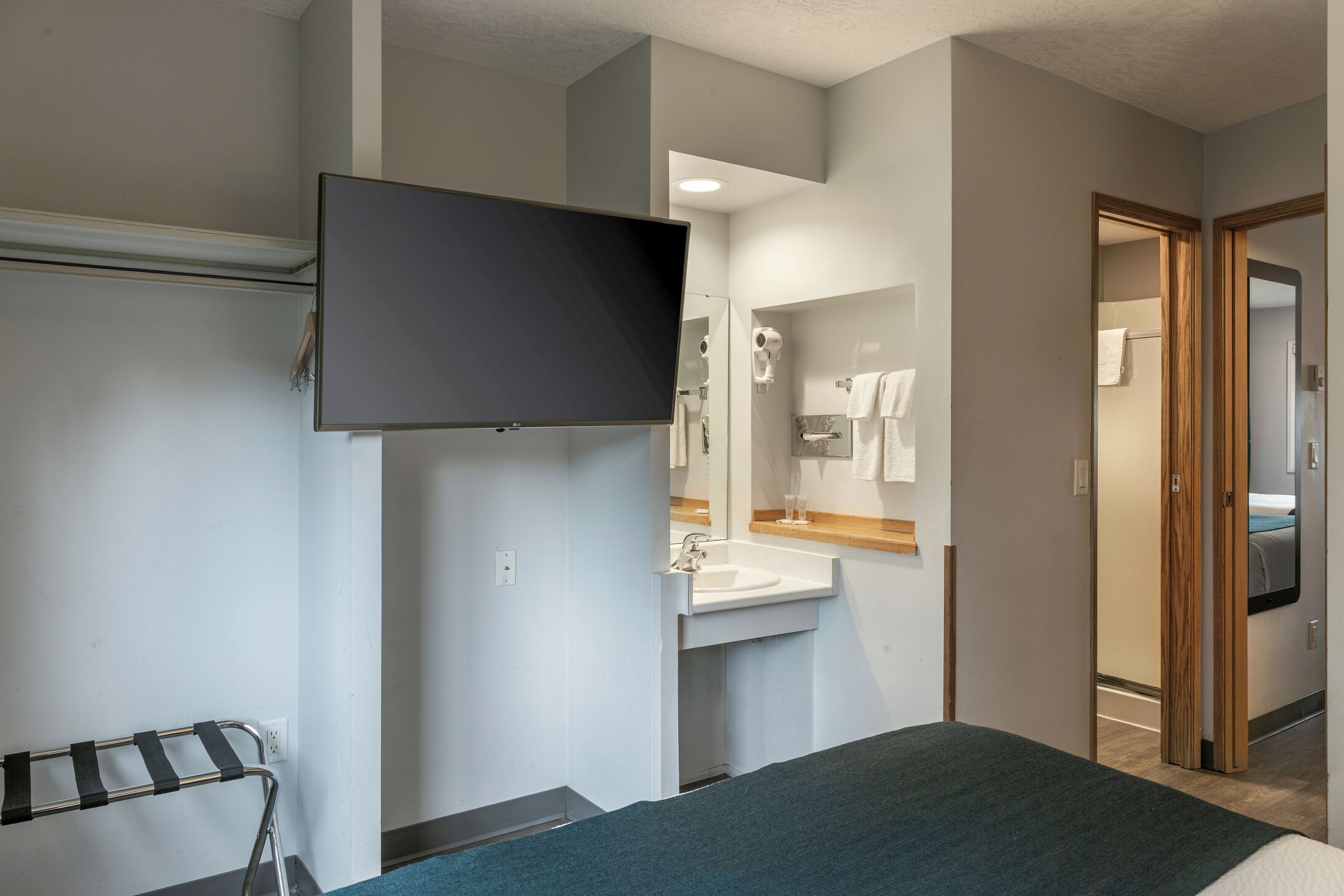 A hotel room with a bed, a TV mounted on the wall, a bathroom with a sink and mirror, and a towel rack.