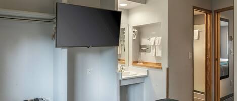 A hotel room with a bed, a TV mounted on the wall, a bathroom with a sink and mirror, and a towel rack.