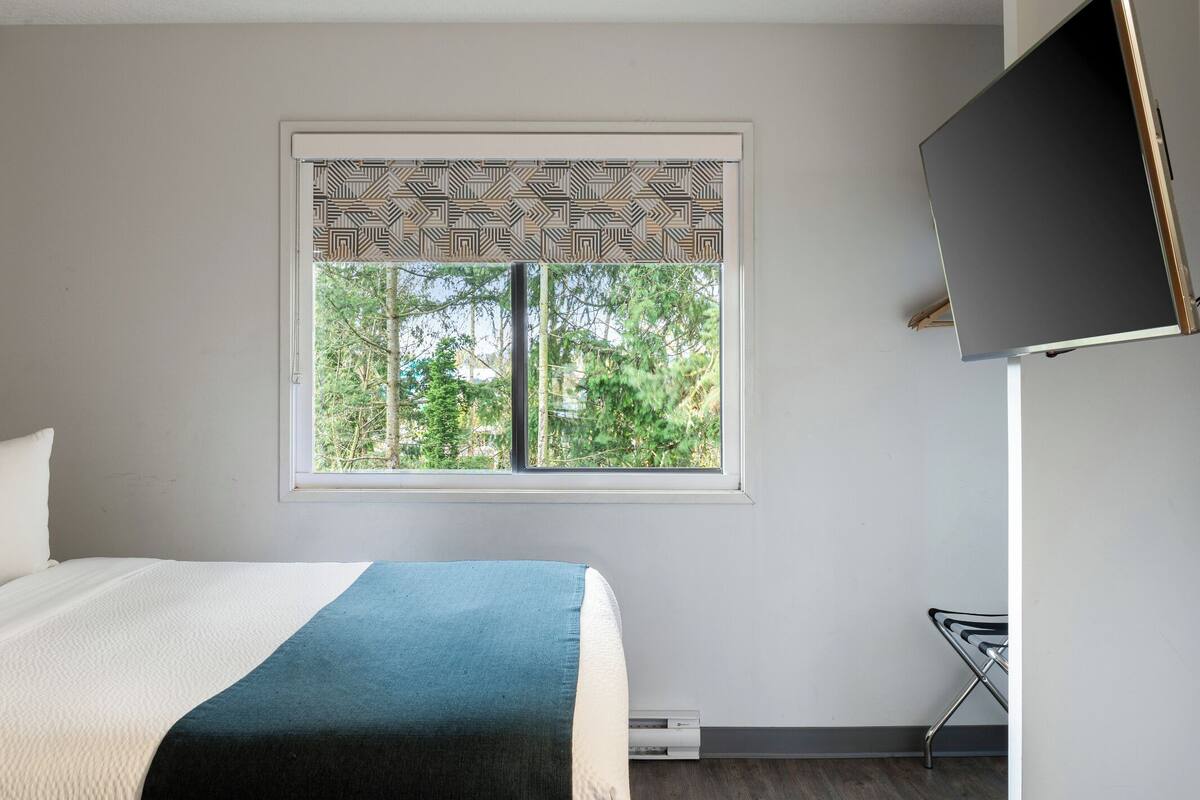 A bedroom with a bed, a window with patterned blinds, a flat-screen TV mounted on the wall, and a chair.