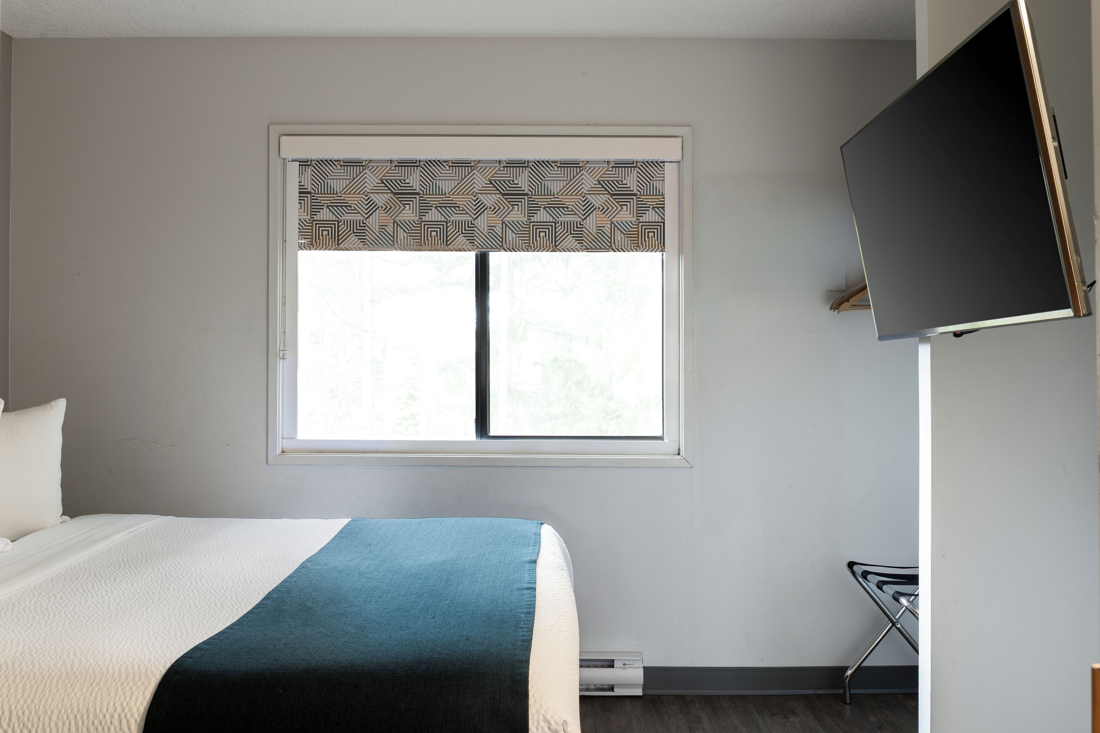 A bedroom with a bed, a window with patterned curtains, a flat-screen TV mounted on the wall, and a radiator.
