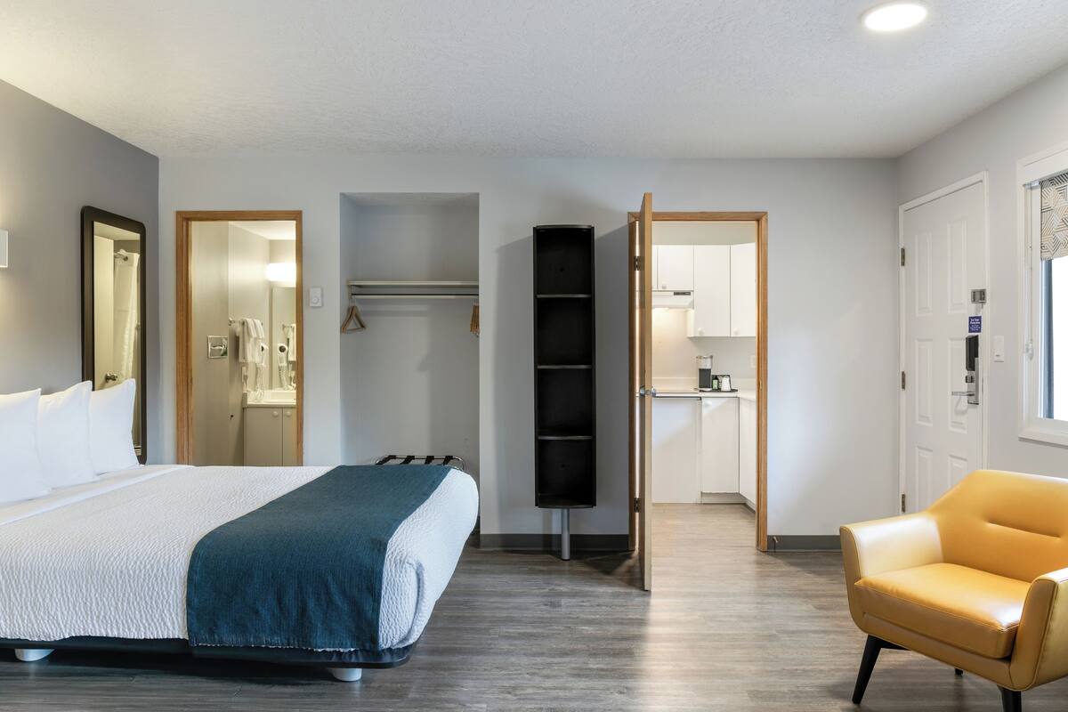 A hotel room with a bed, a chair, a wardrobe, a mirror, and a bathroom visible through an open door.