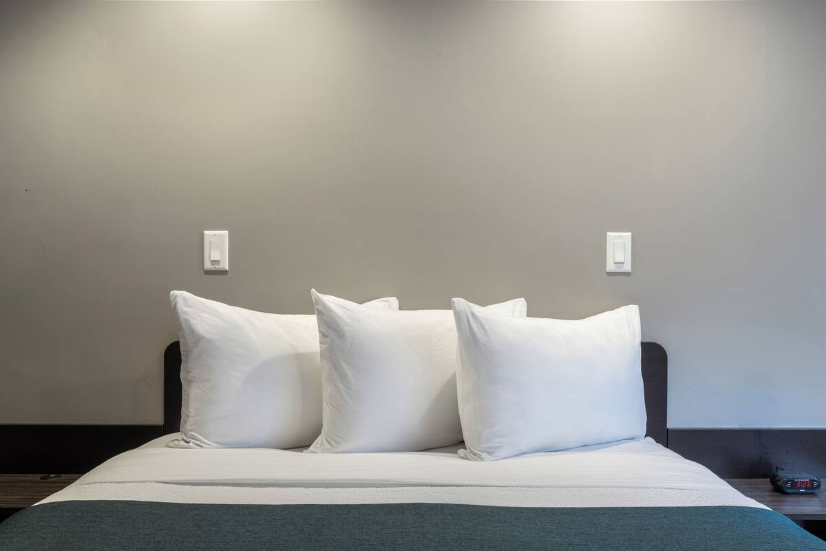 A bed with white bedding and three pillows, a bedside table with a clock, and a wall-mounted light fixture.