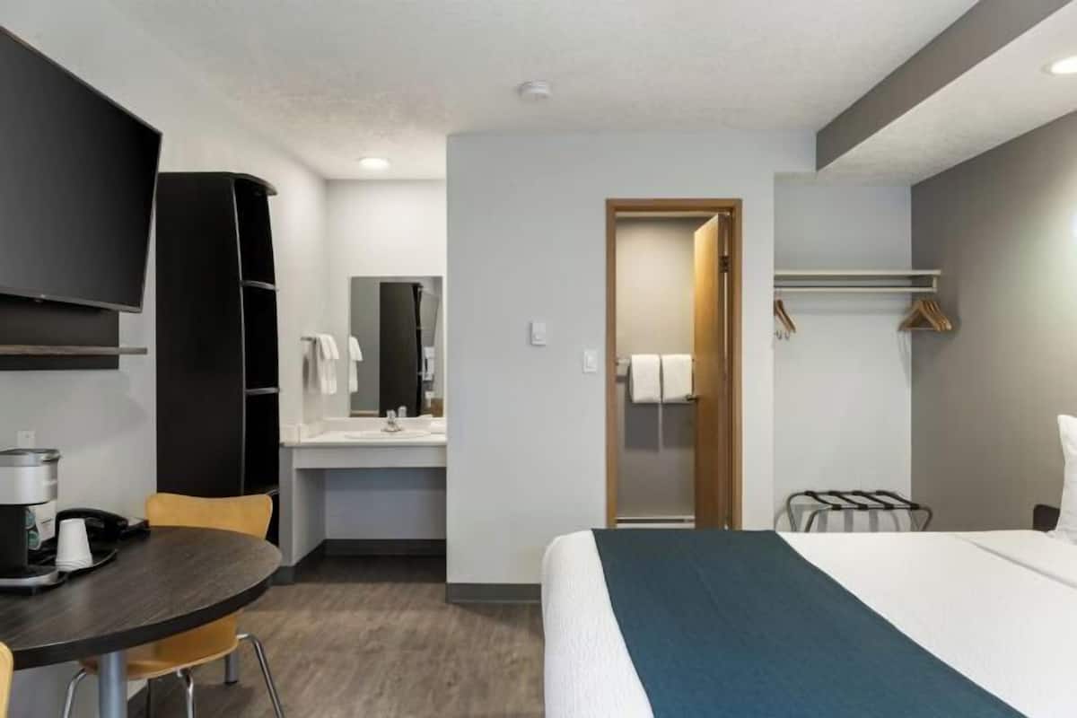 A hotel room with a bed, a desk with a chair, a television, a mirror, a bathroom door, and a coat rack.