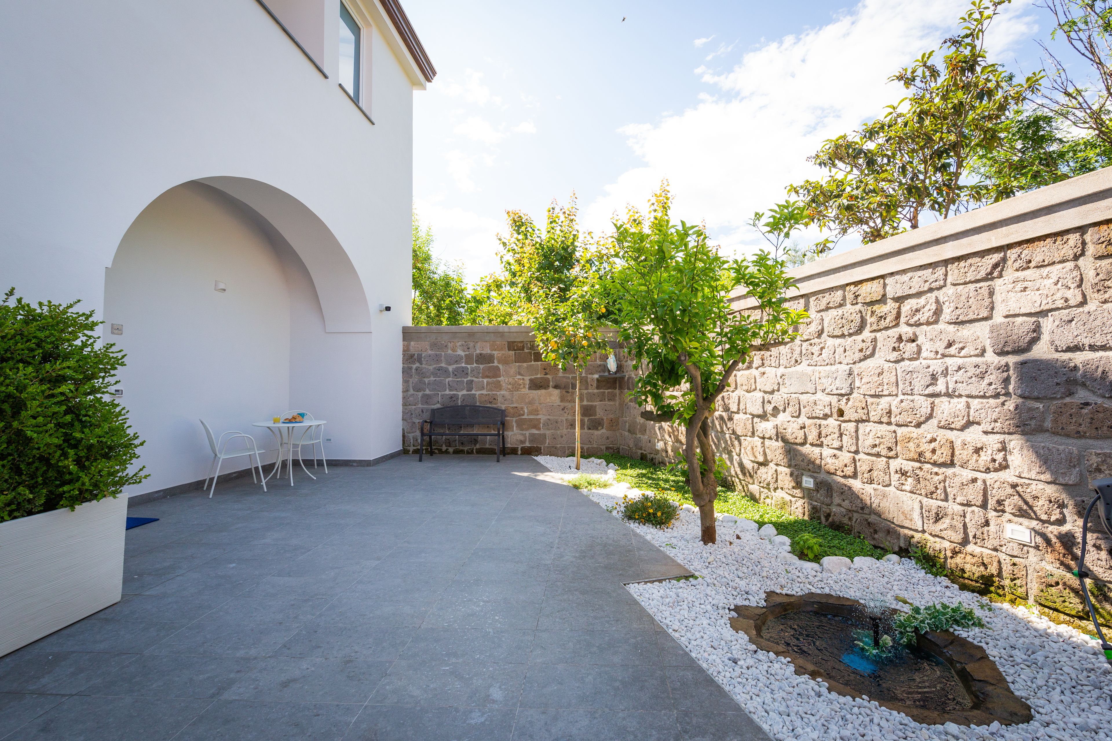A courtyard with a stone wall, a small tree, a white planter, and a seating area with chairs.