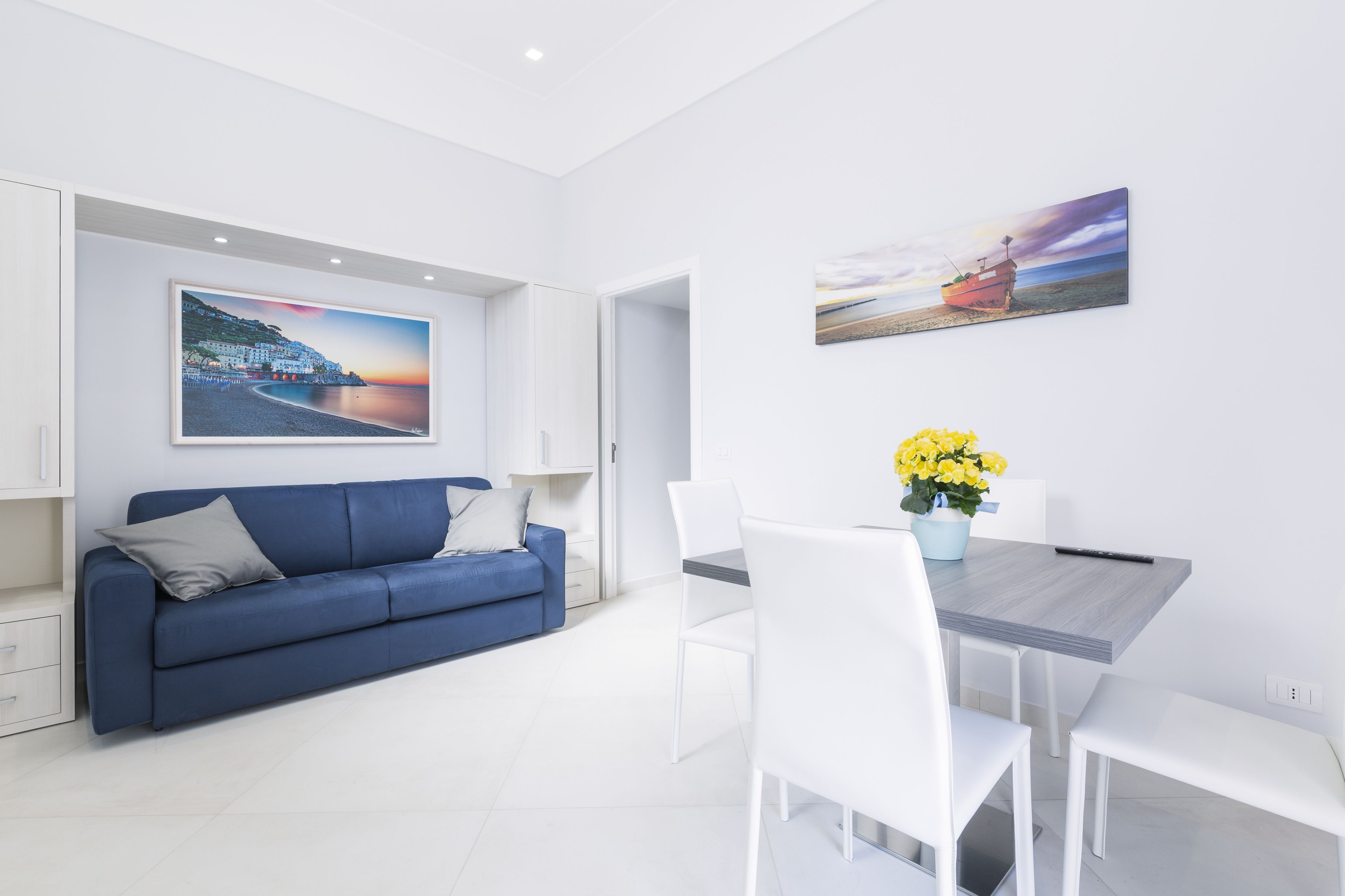 A modern living room with a blue sofa, white dining table, and chairs, a painting of a coastal scene, and a vase with yellow flowers.