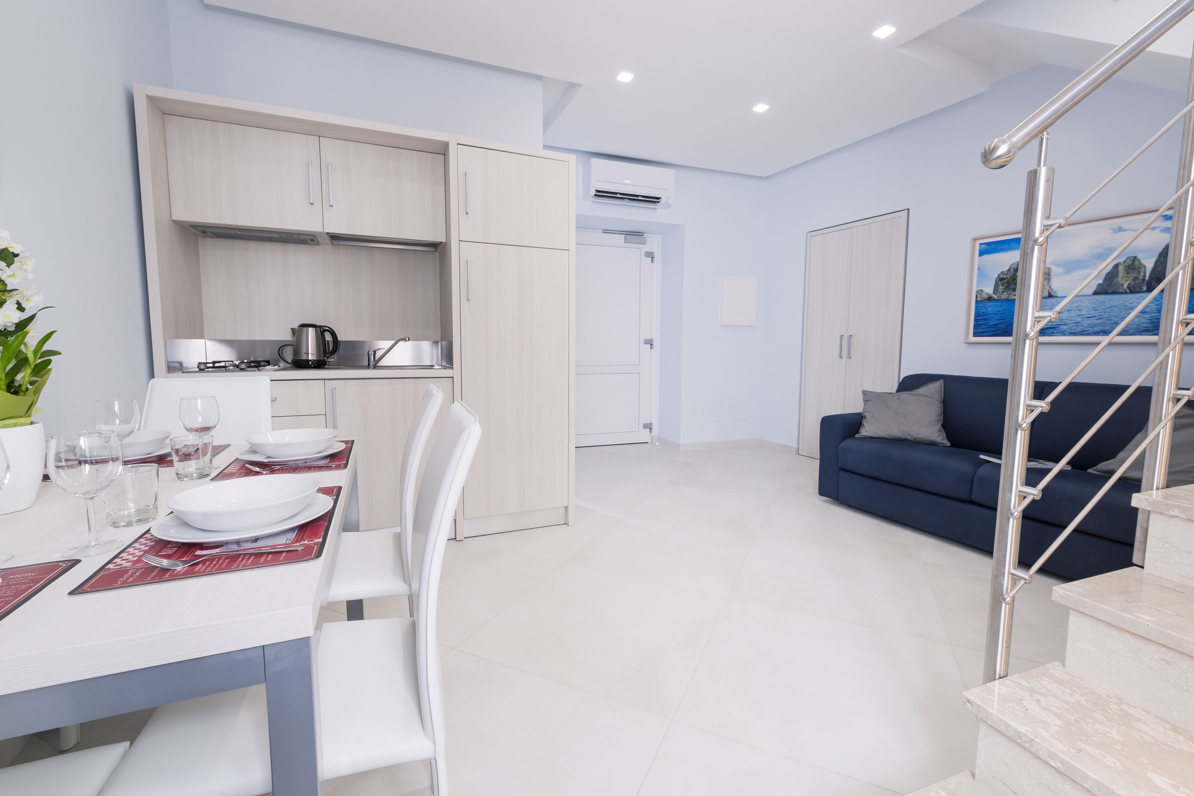 Una cocina moderna con armarios blancos, una mesa de comedor preparada para cuatro personas y una escalera que conduce a una sala de estar con un sofá azul.
