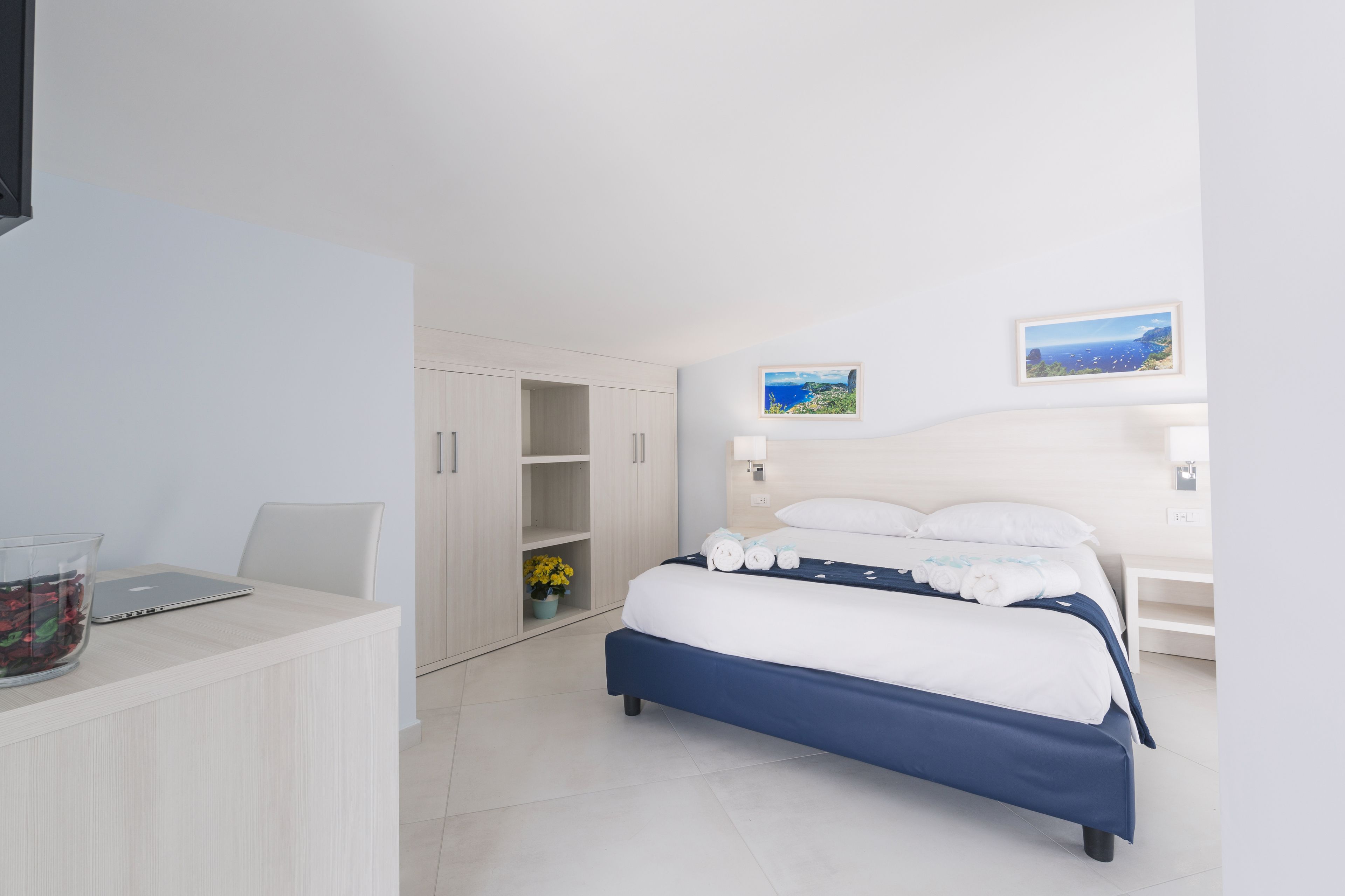 A modern hotel room with a bed, a desk with a laptop, a wardrobe, and a painting on the wall.