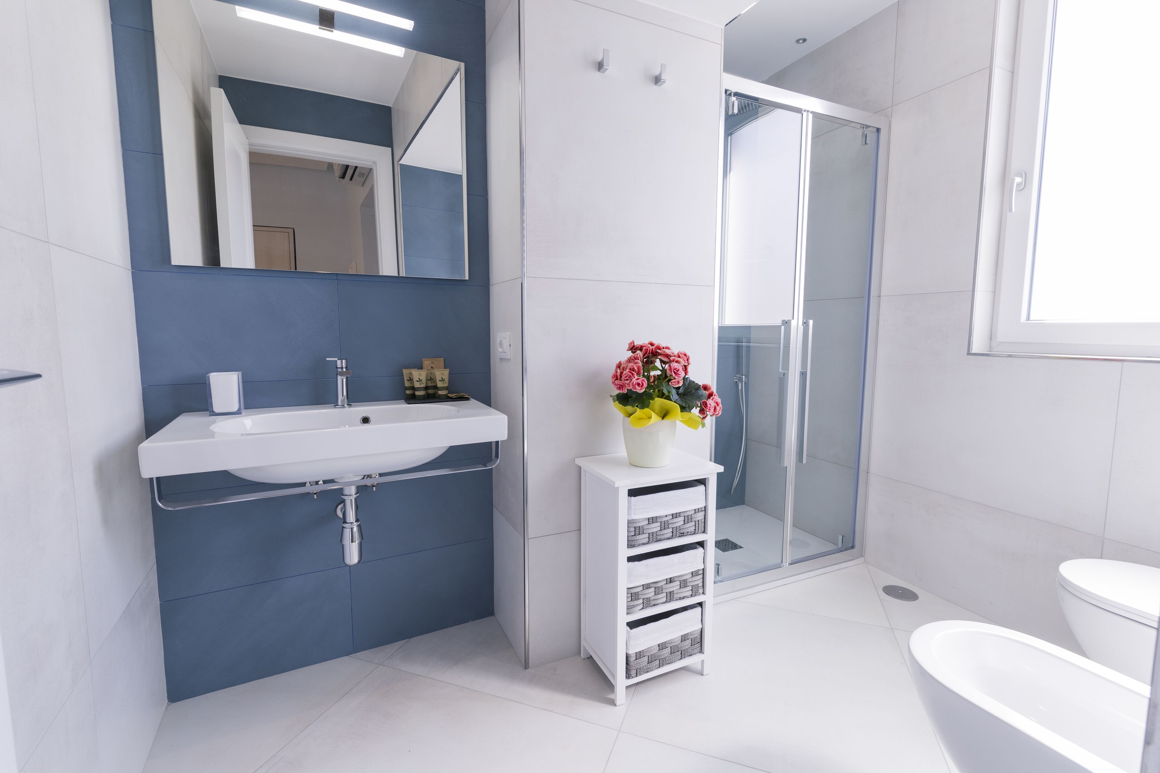 Et moderne badeværelse med en hvid vask, et spejl, et lille hvidt skab med skuffer, en glasbrusekabine og et hvidt toilet.