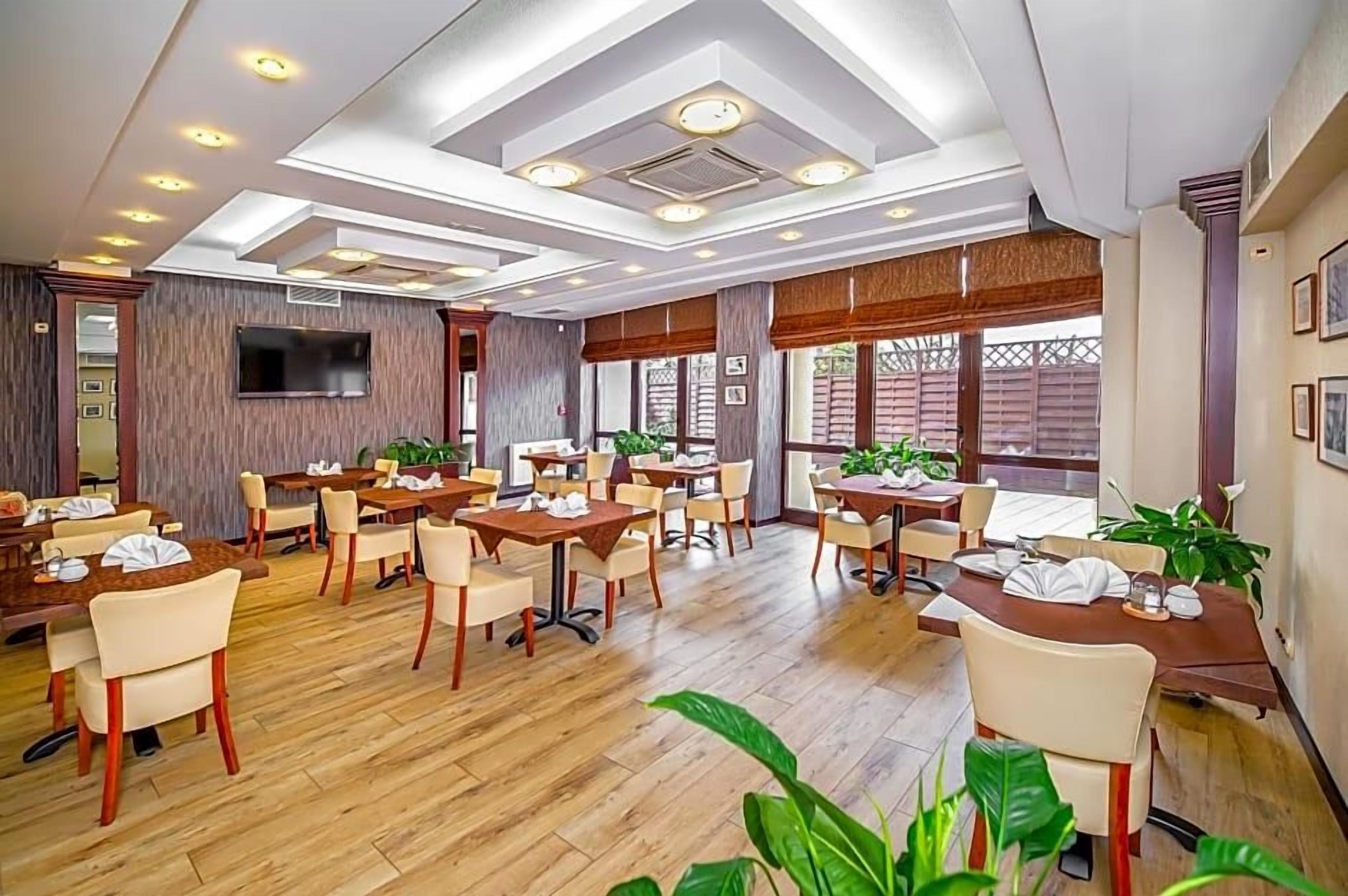 A restaurant with multiple tables and chairs, a flat-screen TV mounted on the wall, and large windows allowing natural light.
