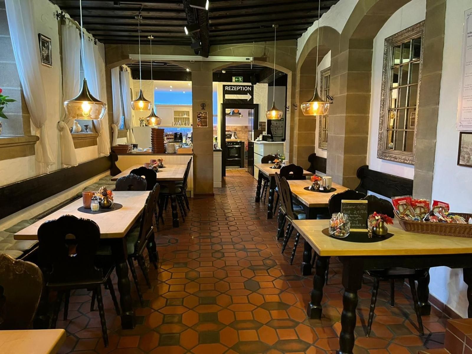 A restaurant interior with hexagonal patterned flooring, wooden tables and chairs, pendant lighting, and a reception area in the background.