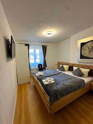 Cama doble con estructura de madera, dos almohadas y una colcha. Hay una mesita de noche con una lámpara. La habitación tiene paredes blancas y suelo de madera.