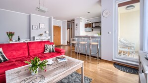A modern apartment with a red sofa, a wooden coffee table, a kitchen with a bar, and a large window with wicker furniture.