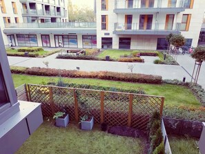 Ein modernes Gebäude mit mehreren Balkonen, einem angelegten Innenhof mit ordentlich geschnittenen Hecken und einem umzäunten Bereich mit Kübelpflanzen.