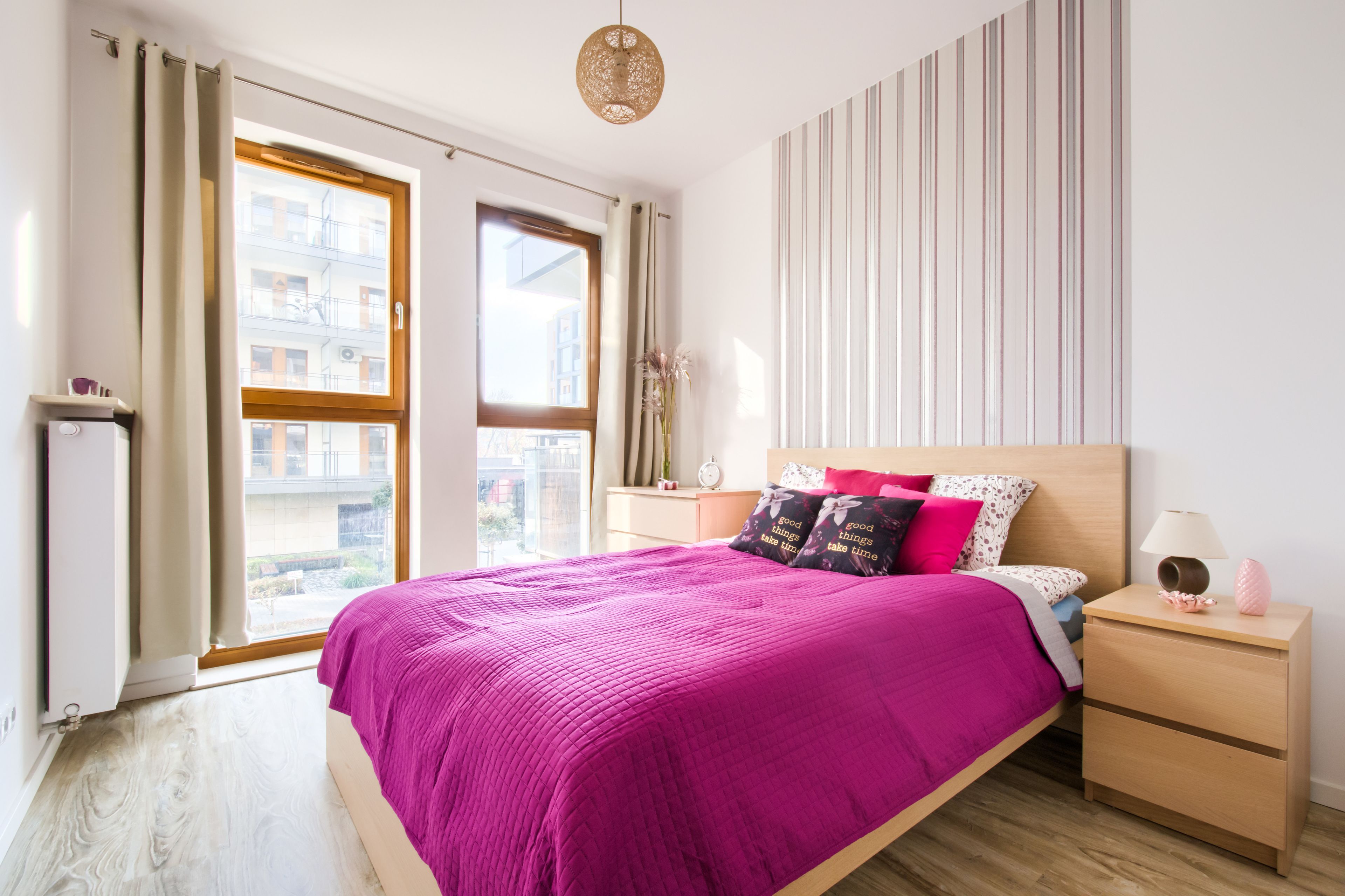 A bedroom with a large bed, a bedside table, a nightstand, a window with curtains, and a view of buildings outside.