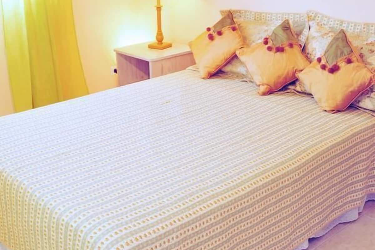 A neatly made bed with a patterned bedspread and matching pillows, a bedside lamp, and a framed picture on the wall.