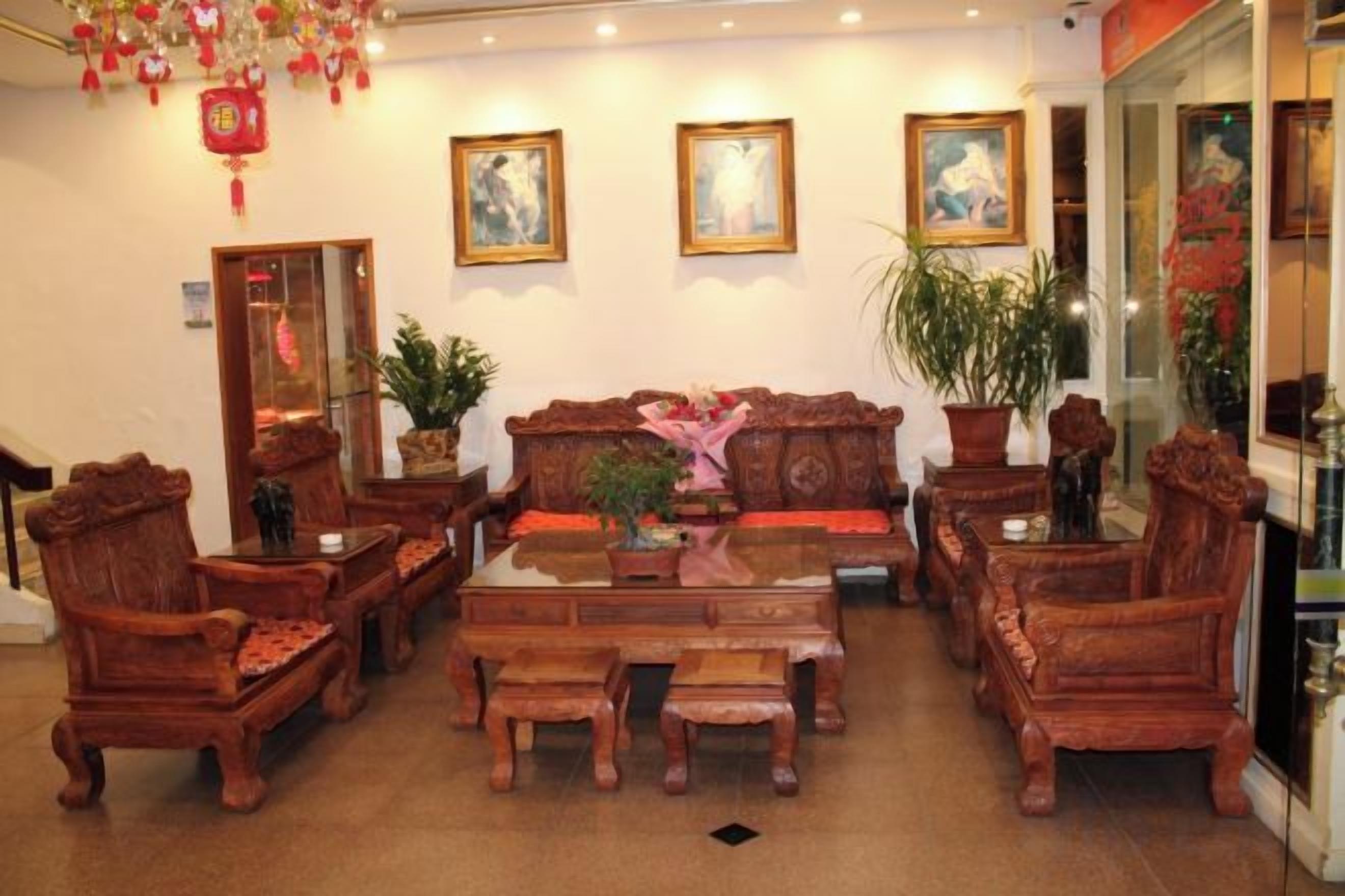 A traditional-style living room with wooden furniture, including sofas and armchairs, a coffee table, and a side table. There are several potted plants and framed artworks on the walls.