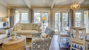 A spacious living room with a beige sofa, patterned armchairs, a round wicker coffee table, and a dining area with a white table and chairs. Large windows provide natural light and views of the outdoors. - Charming Ocean view Retreat! Newly Remodeled (Hilton Head Island)