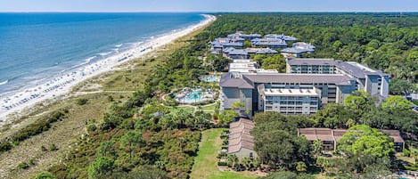 Vista aérea de un complejo turístico frente al mar, con playa, varias construcciones y una piscina.
