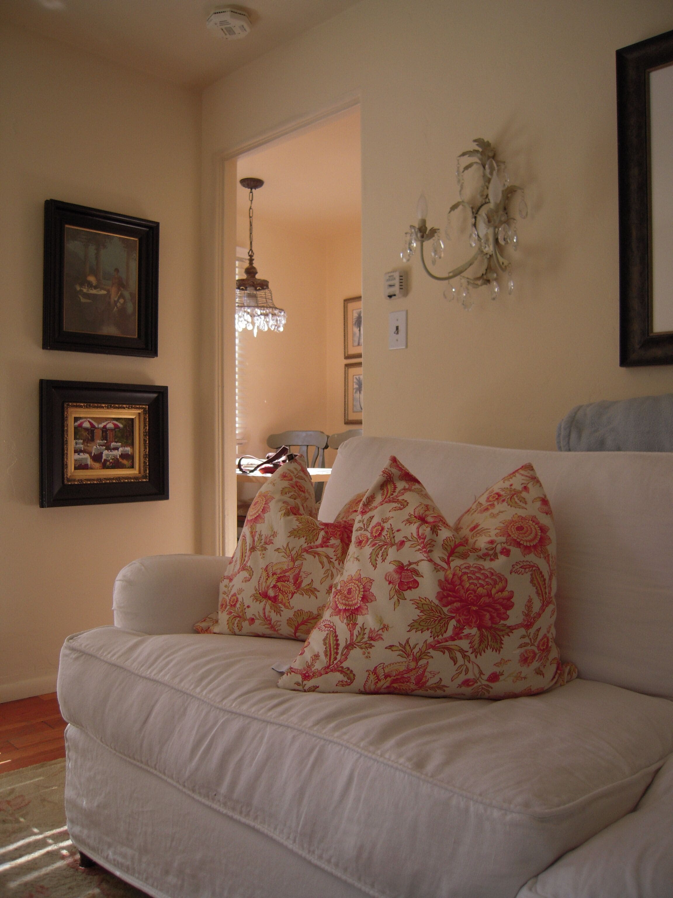 A living room with a white sofa, floral cushions, a chandelier, and framed artwork on the walls.