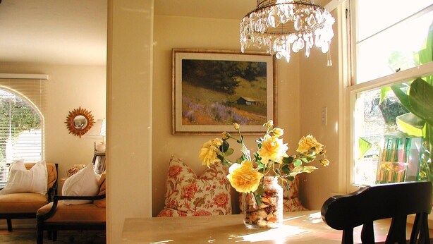 Een eethoek met een tafel, stoelen, een vaas met gele bloemen, een schilderij aan de muur en een kroonluchter.