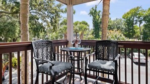Un balcon avec deux chaises et une table, donnant sur une rue bordée de voitures garées et d’arbres.