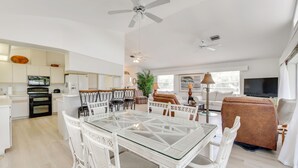 Amplia sala de estar con una mesa de comedor de vidrio rodeada de sillas de mimbre blanco, un sofá marrón y un televisor.