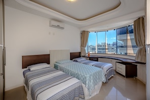 A hotel room with two beds, a large window with curtains, a built-in desk, and an air conditioning unit.