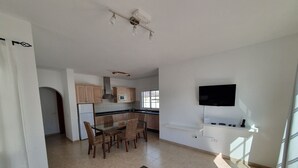 A modern kitchen with wooden cabinets, a dining area with a glass table, and a TV mounted on the wall.