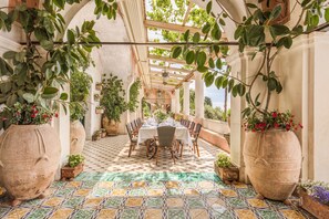 A covered outdoor dining area with a tiled floor, a table set for a meal, and large potted plants.