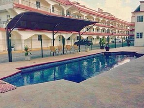A swimming pool with lounge chairs and a covered seating area, surrounded by a multi-story building with balconies.