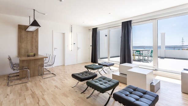 A modern, well-lit room with a wooden floor, a dining area with chairs and a table, and a large window offering a view of the sea. - Apartment Villa Mila (Kaštel Novi)