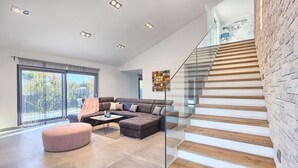 A modern living room with a sectional sofa, a glass staircase, and a large window with a view of trees. - Villa Fortuna - in historic town Buje with 4 bedrooms and a pool, near the beach by MyWaycation (Kaštel)