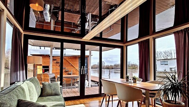 Een modern interieur met een houten vloer, een groene bank, een eettafel met stoelen en grote ramen met uitzicht op een waterpartij.