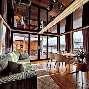 Un intérieur moderne avec un plancher en bois, un canapé vert, une table à manger avec des chaises et de grandes fenêtres donnant sur un plan d’eau.
