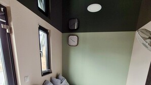 Een kleine slaapkamer met een bed, kussens, een klok en een raam.