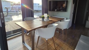 Een modern interieur met een houten eettafel, witte stoelen, een flatscreen-tv en uitzicht op een boot door grote ramen.