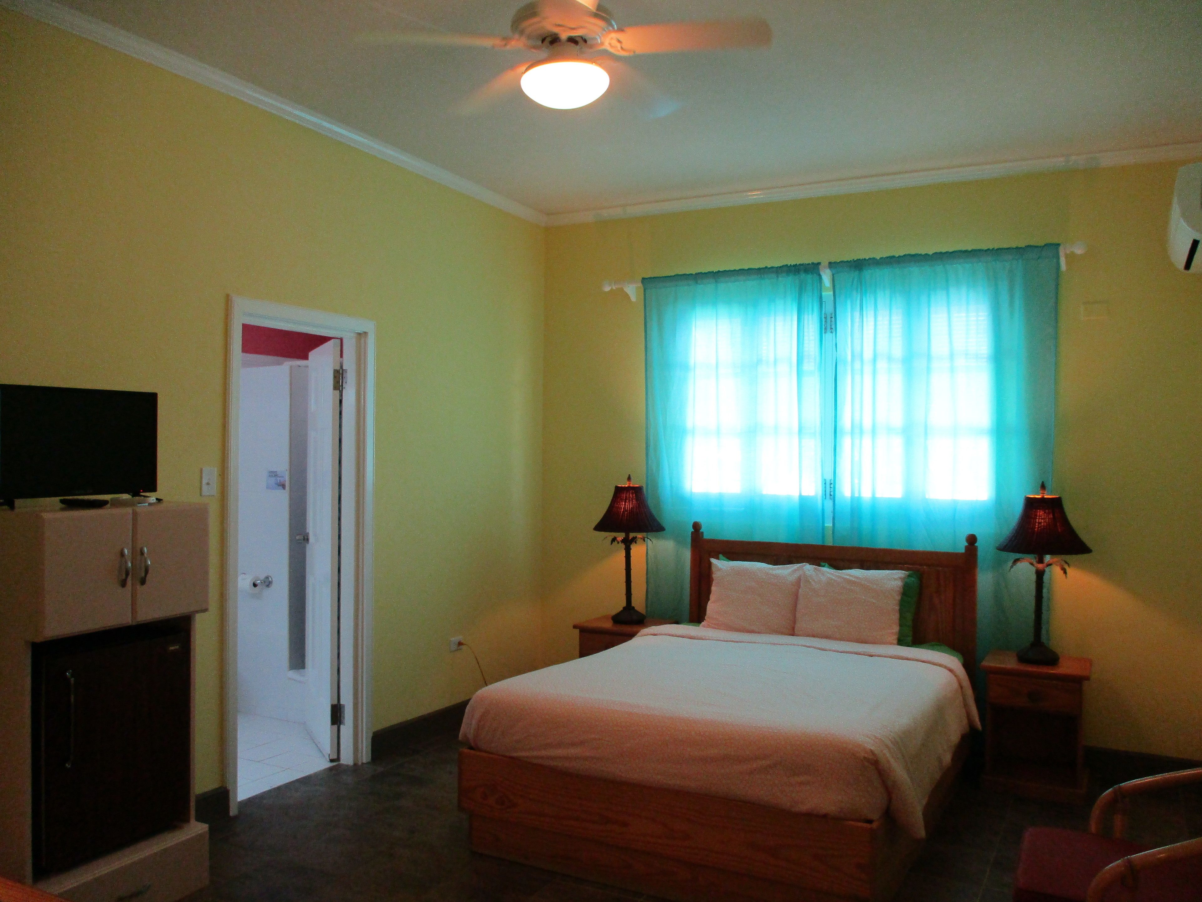 A bedroom with a bed, two bedside lamps, a ceiling fan, a television on a cabinet, and a window with curtains.